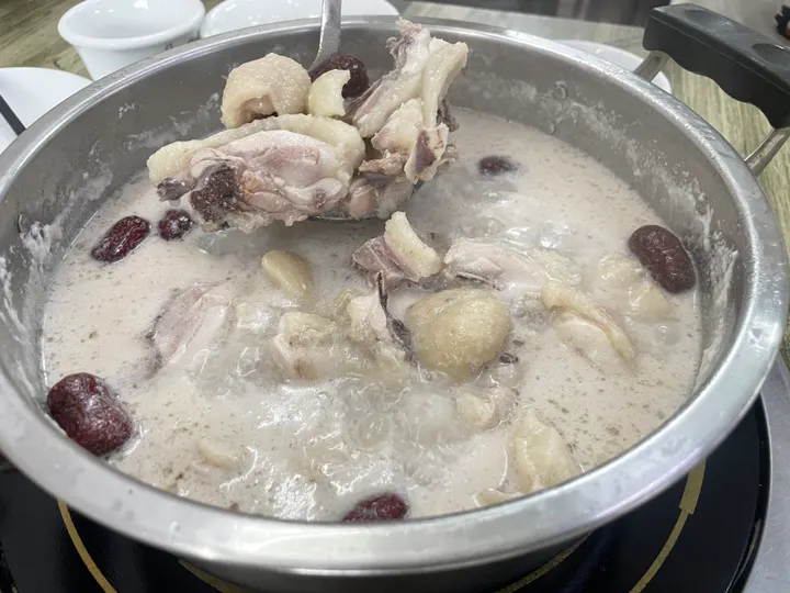 An original photograph of bone-in chicken pieces and red dates boiling in a metal pot of coconut broth, with a ladle lifting several pieces