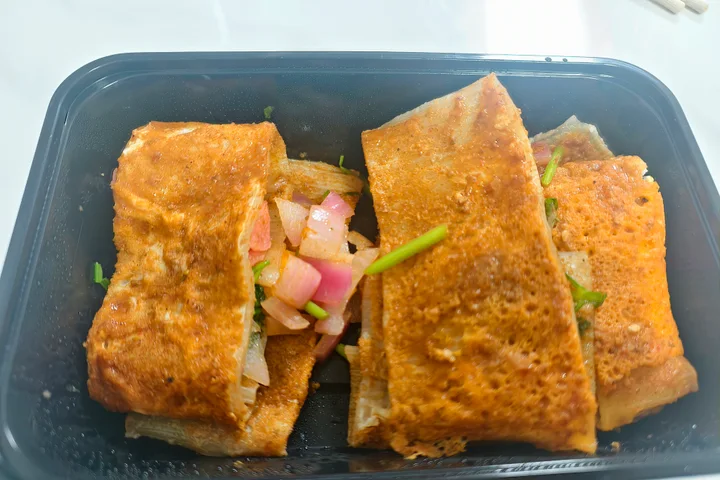 An original photograph of folded grilled cold noodle pieces with diced red onion, coriander and sausage in a black takeaway tray