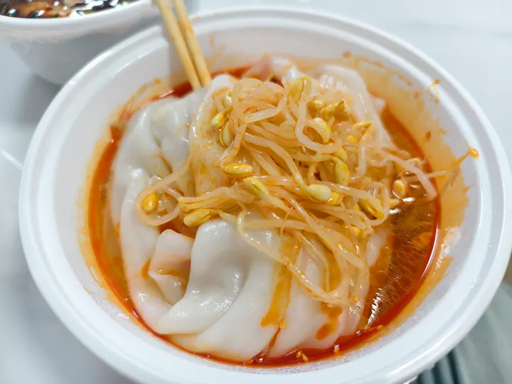 An original photograph of broad hot rice pi sheets with soybean sprouts and red chilli seasoning in a white bowl