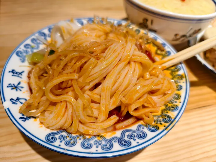An original photograph of cold-dressed liangpi ribbons coated with red chilli seasoning on a blue-and-white plate