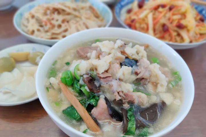 A finished bowl of Xi’an yangrou paomo with torn bread, sliced lamb, glass noodles, wood-ear mushroom and herbs, with pickled garlic and cold dishes behind it